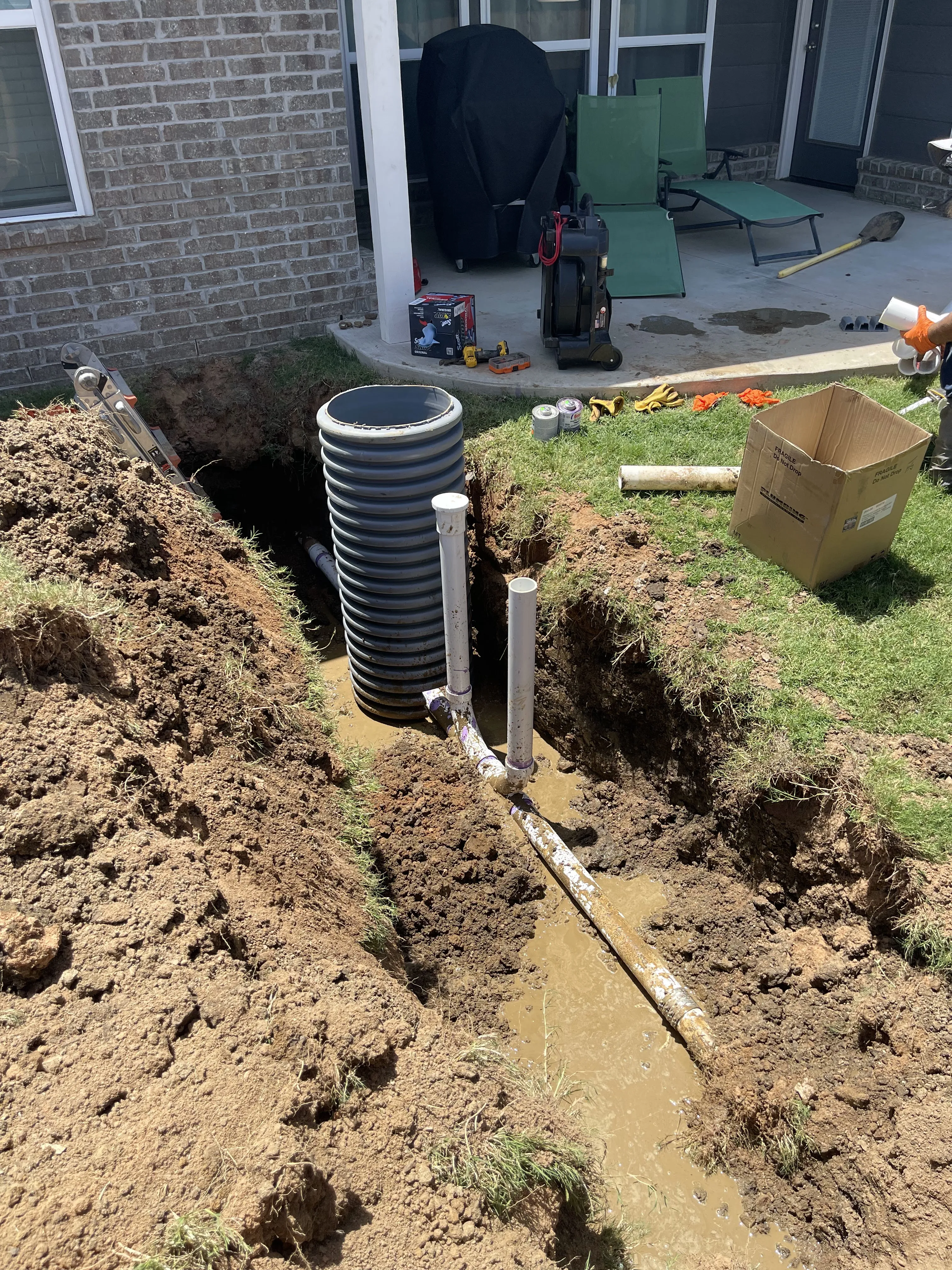 A corrugated riser and PVC sewer line exposed in a trench beside a brick house